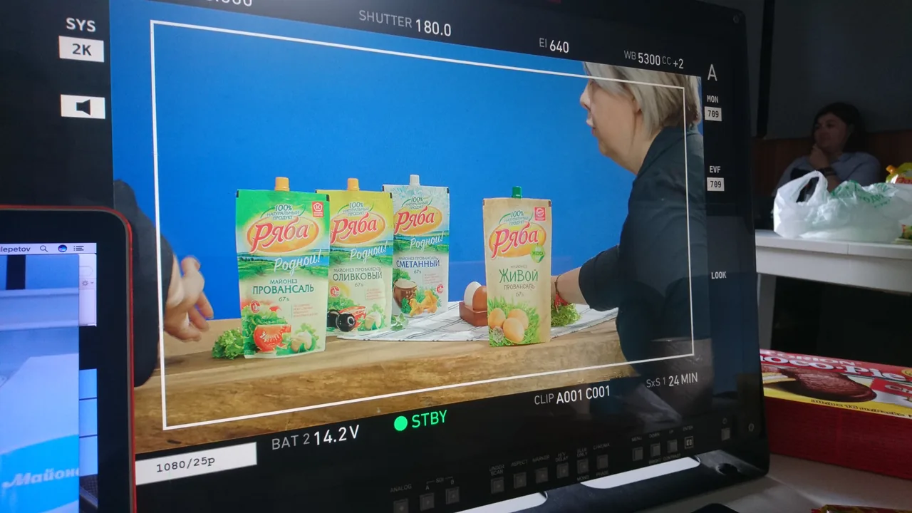 Крупный план монитора: упаковки «Ряба», продукты и человек, поправляющий композицию.