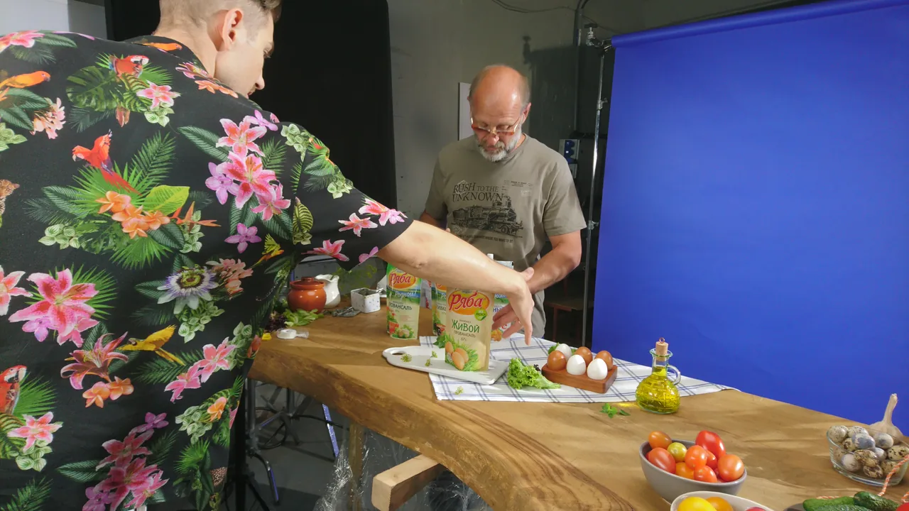 Двое людей выставляют упаковки «Ряба» и продукты на столе перед синим фоном.